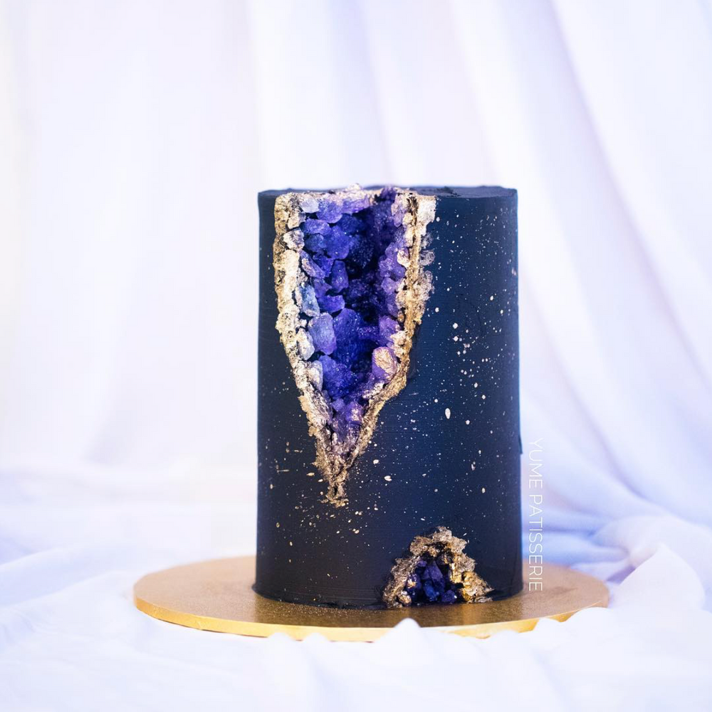 A tall and slim cake, this cake is covered in a dark bluish purple fondant. The fondant has speckles of gold splattered on it that resemble stars. A chunk of the cake has been cut out adn replaced with edible purple rock candy that resemble Amethyst gems, so the cake looks like a geode.