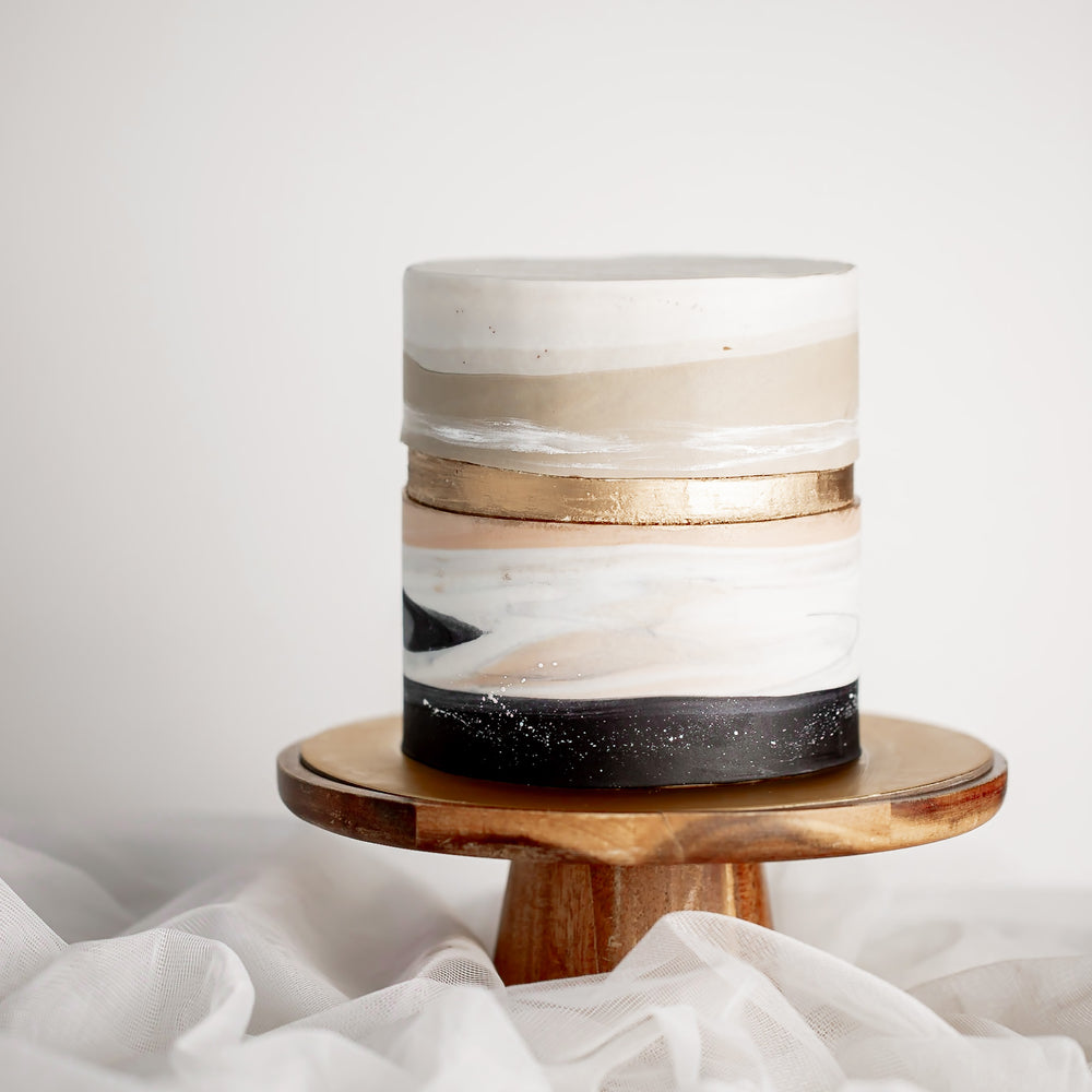 A cake with a marbled white and beige fondant base that resembles sand. The bottom of the cake has black fondant that resembles a stone floor, and the middle of the cake has a golden faultline.
