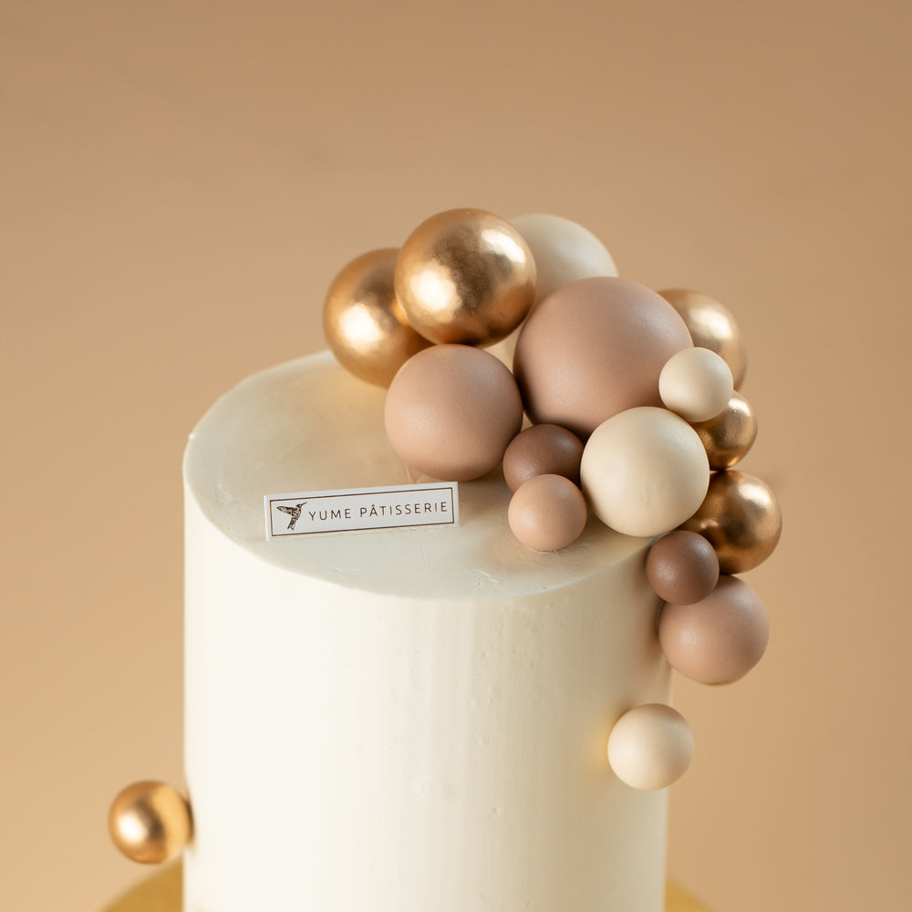 A tall and slim cake with a white buttercream base. The cake has numerous shiny and matte round ornaments in gold and nude colours on the top and bottom of the cake.