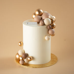 
                  
                    Load image into Gallery viewer, A tall and slim cake with a white buttercream base. The cake has numerous shiny and matte round ornaments in gold and nude colours on the top and bottom of the cake.
                  
                