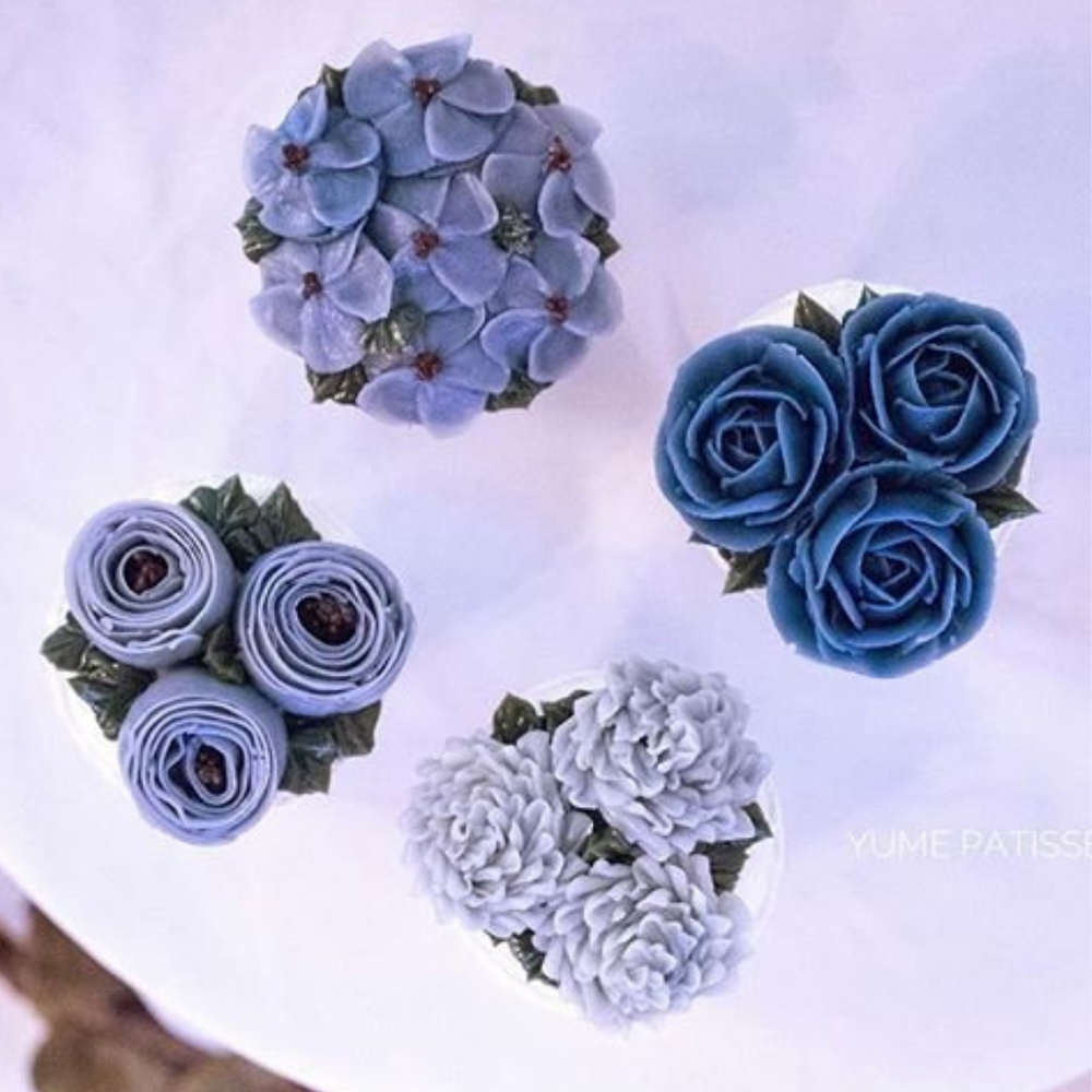 Four cupcakes, all in assorted shades of blue. The cupcakes have assorted buttercream flowers on them, and look incredibly realistic