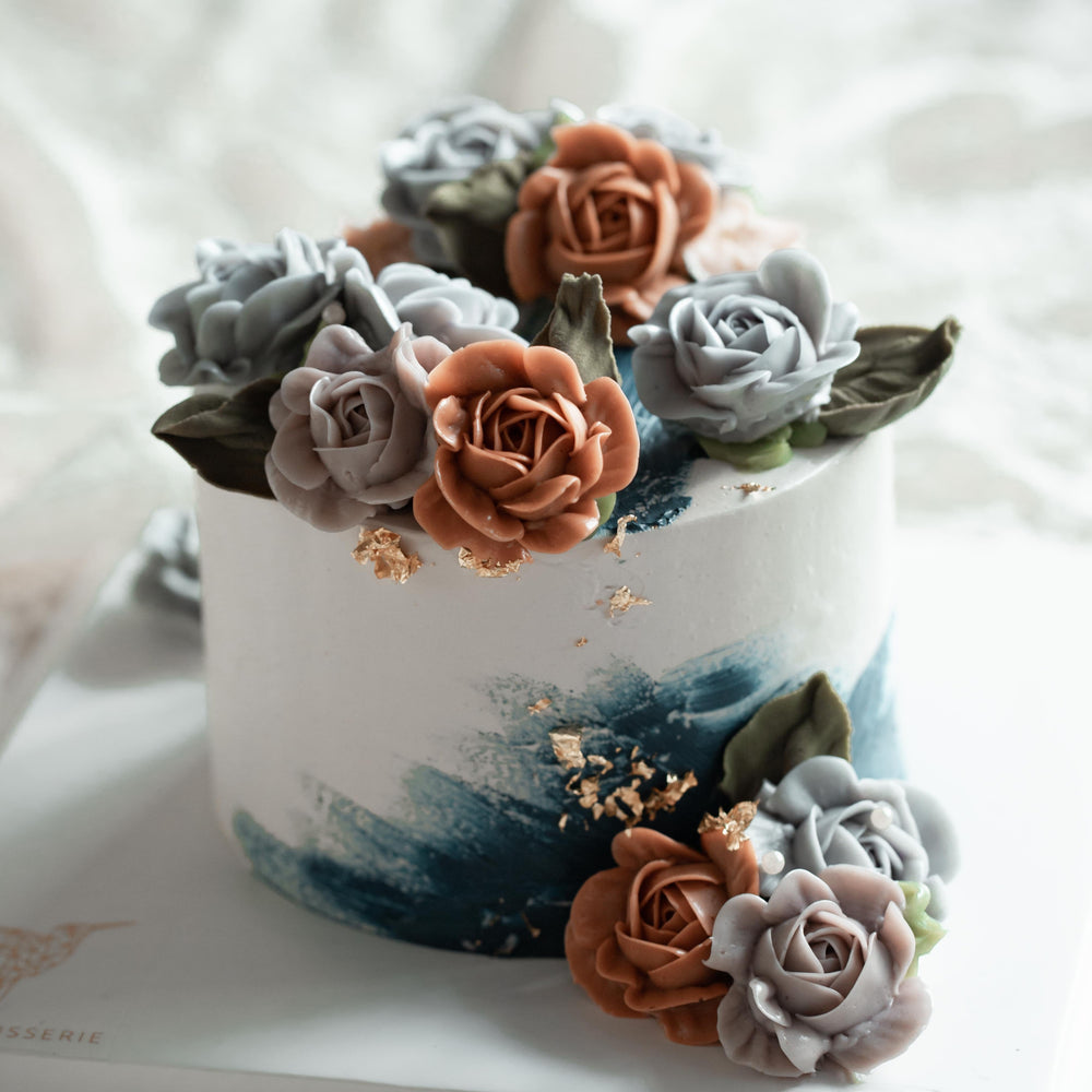 A white buttercream cake with accents of dark blue buttercream. The cake has muted orange, purple and blue buttercream flowers, and bits of gold leaf.