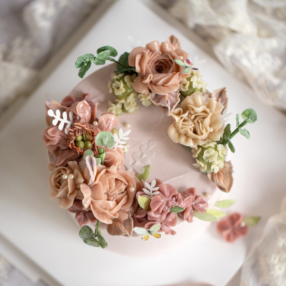 A light pink. buttercream base, with pink, beige and ivory buttercream flowers on top in a wreath style.