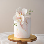 A tall cake with light pink fondant finishing, with streaks of pink and gold leaf marbled into the fondant. The top of the cake has two white edible sugar orchids.