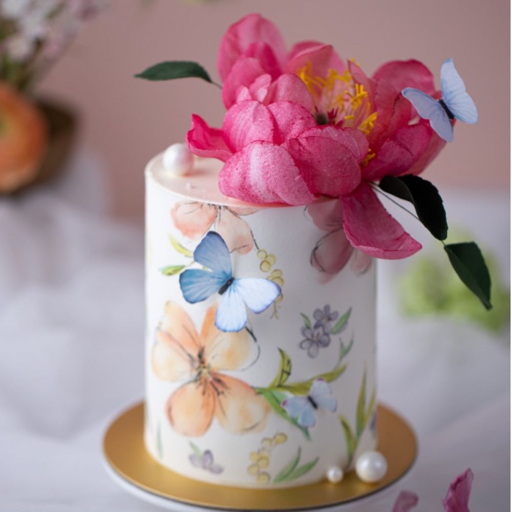 A small but tall cake with a white fondant base, and watercolour style flowers on an edible icing sheet wrapped around the cake. There are edible wafer butterflies on the cake, along with a realistic edible wafer peopny on top. The peony is a deep shade of pink, and looks very detailed. There are edible sugar pearls scattered around the cake.