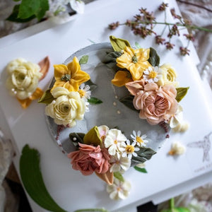 
                  
                    Load image into Gallery viewer, A dusty, warm grey buttercream cake with bright pops of yellow, ivory and coral buttercream flowers on top in a wreath style. The cake also has edible sugar pearls delicately scattered on top and around it.
                  
                