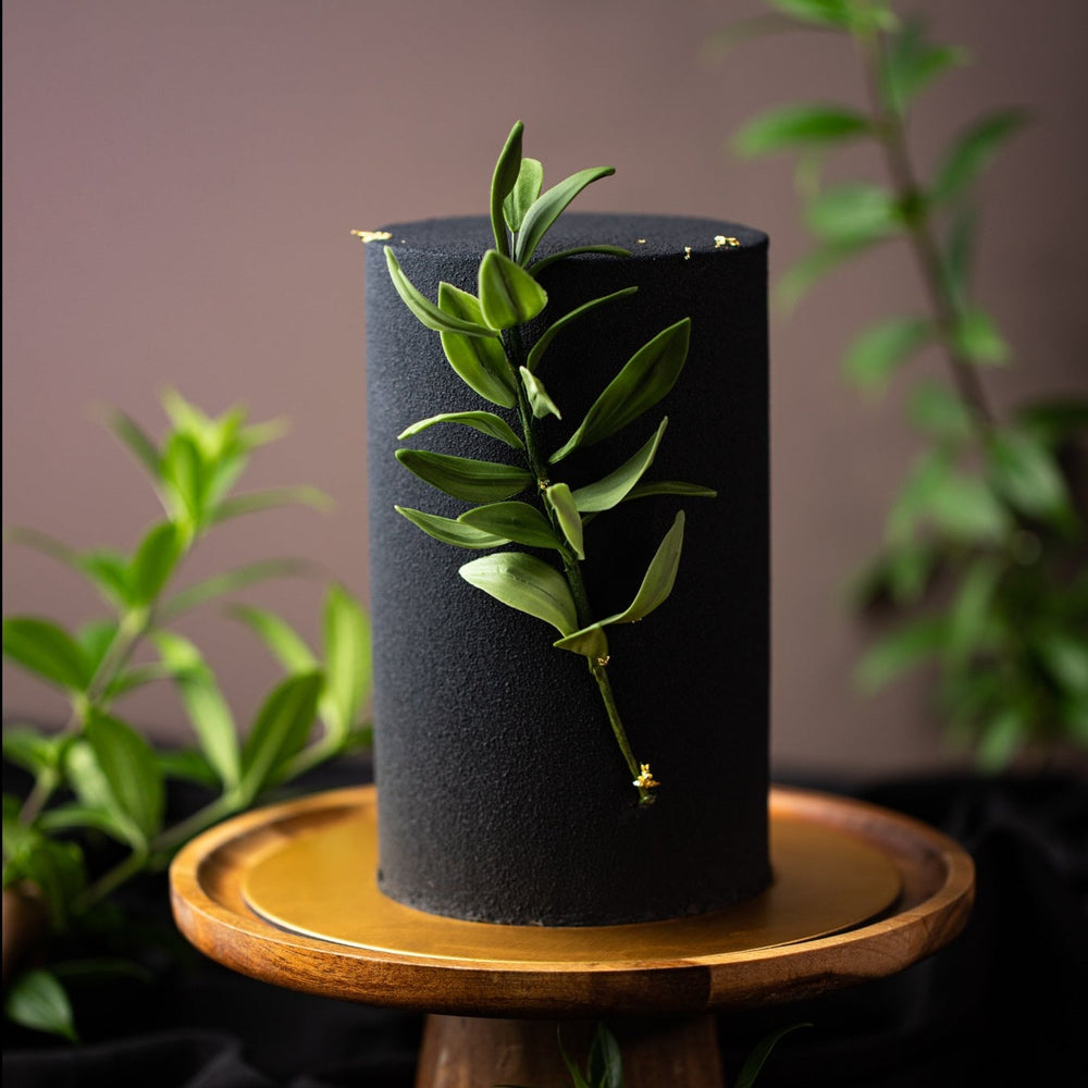 A tall and slim cake that has beenm sprayed with black chocolate velour spray. The cake has edible green sugar leaves on the front, and it looks incredibly realistic.