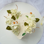 A heart shaped cake with white buttercream base, and ivory buttercream flowers on top of the cake.