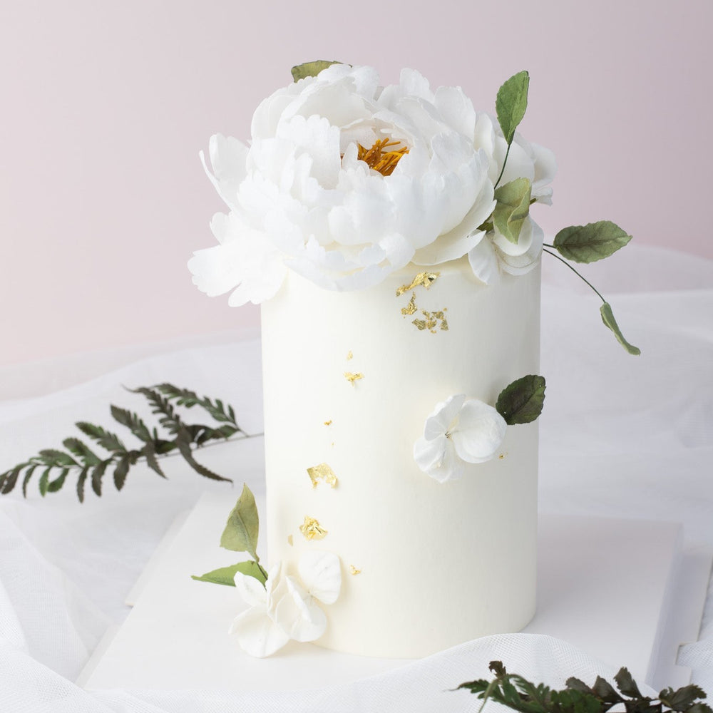 A small but tall cake with a white fondant finish. The cake has bits of edible gold leaf delicately scattered around the side. On top of the cake, there is a white edible wafer peony, along with some green edible wafer leaves. The peony is incredibly realistic, and even has a yellow middle that resembles pistils.