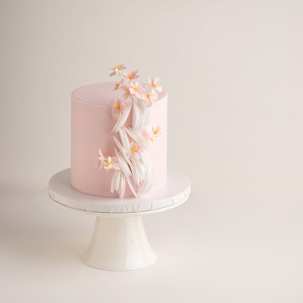 A light ponk fondant base on a tall cake, with delicate white edible wafer paper scallops cascading down one side of the cake. There are small edible wafer sakura flowers on the cake, giving it a Japanese vibe.
