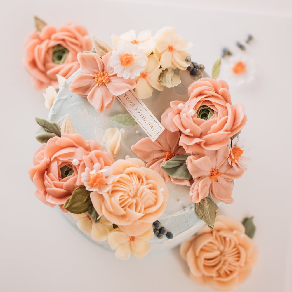 A light, dusty blue buttercream cake with pink and coral buttercream flowers on top, along with green edible wafer leaves. The flowers are assorted and realistic.