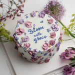 White buttercream cake with light purple and pink palette knife buttercream flowers all over the cake. "Bloom with grace" is piped in cursive buttercream in the middle of the cake.