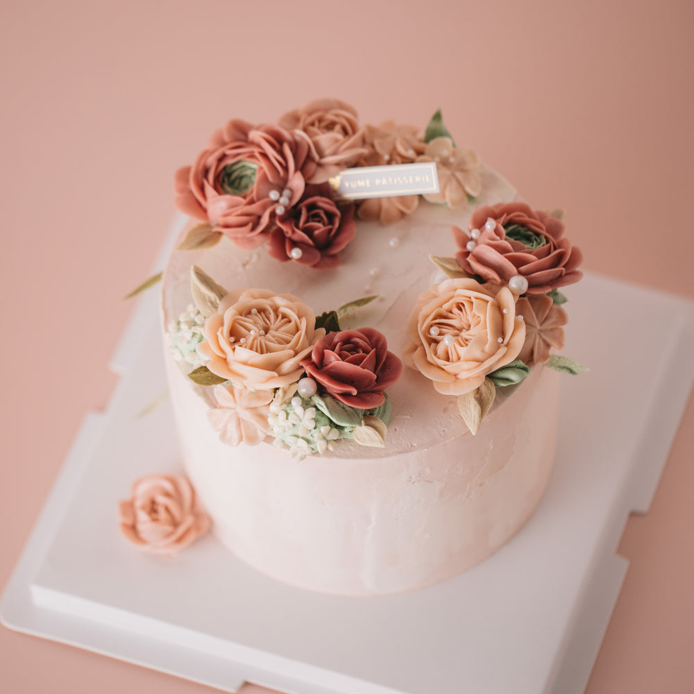 Light pink buttercream cake with red, pink and blush buttercream flowers on top in a wreath style. There are edible sugar pearls scattered on top.
