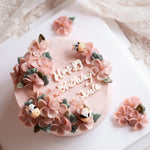 A light pink buttercream cake with pink buttercream flowers on top, decorated with little fondant bees. "Happy Birthday" is written in buttercream cursive in the middle of the cake.