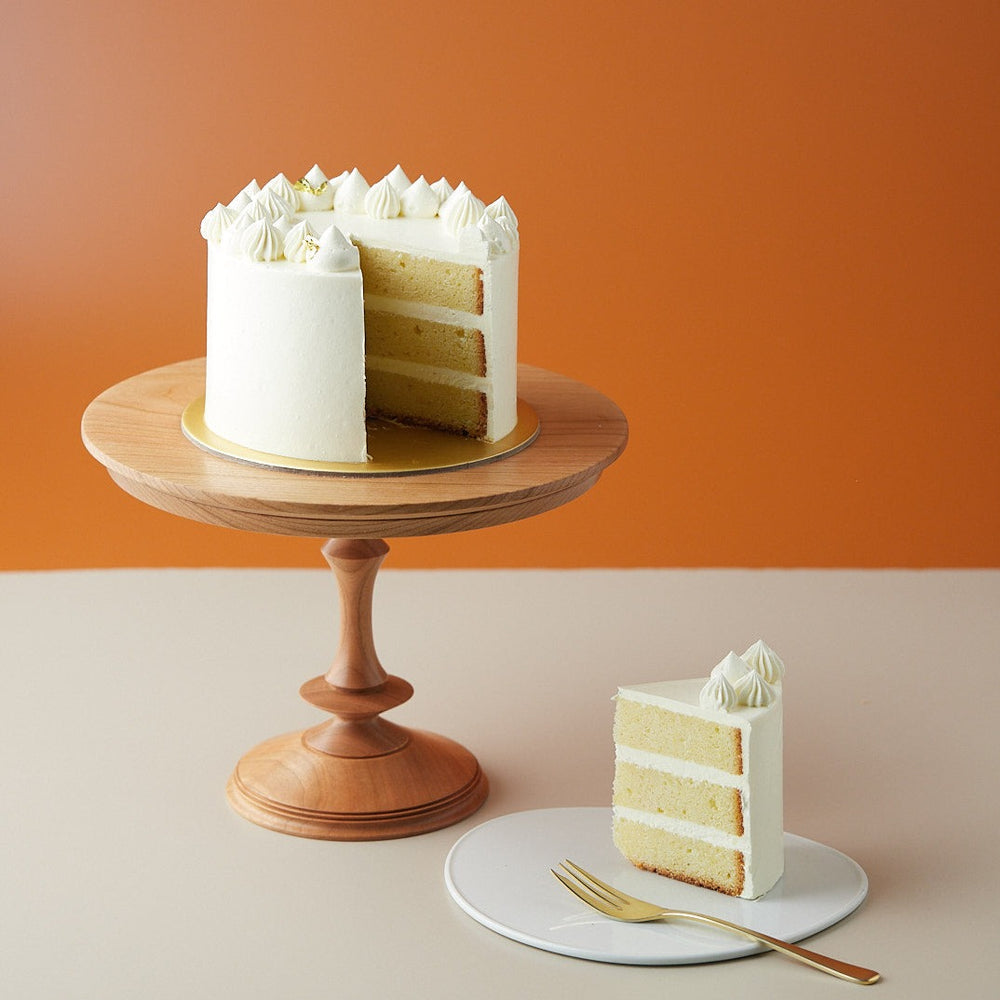 A lemon cake with a slice cut out to show the cross section. The cross section slice is neat and clean, and shows a light yellow sponge that has a tight crumb. The cake looks moist and delicious. The cake has a white buttercream coating.