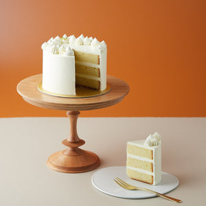 
                  
                    Load image into Gallery viewer, A lemon cake with a slice cut out to show the cross section. The cross section slice is neat and clean, and shows a light yellow sponge that has a tight crumb. The cake looks moist and delicious. The cake has a white buttercream coating.
                  
                