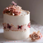 A white buttercream cake base, with a pink faultline in the middle of it. There is gold leaf carefully painted at the edge of the faultline, and the top of the cake. There are pink edible wafer butterflies on the pink faultline. The top of the cake has hand piped buttercream roses in various shades of pink and blush.