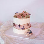 A white buttercream faultline cake, where the "inner" part is a light shade of blush. There are pink and purple edible wafer butterflies in the middle, and the edges of the faultline are painted with gold luster dust. The top of the cake is covered in many hand piped pink buttercream roses.