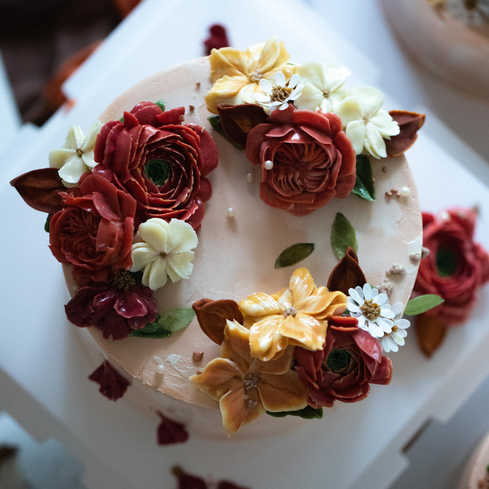 Blush coloured buttercream cake with dark red, orange and white buttercream flowers in assorted shapes on top.