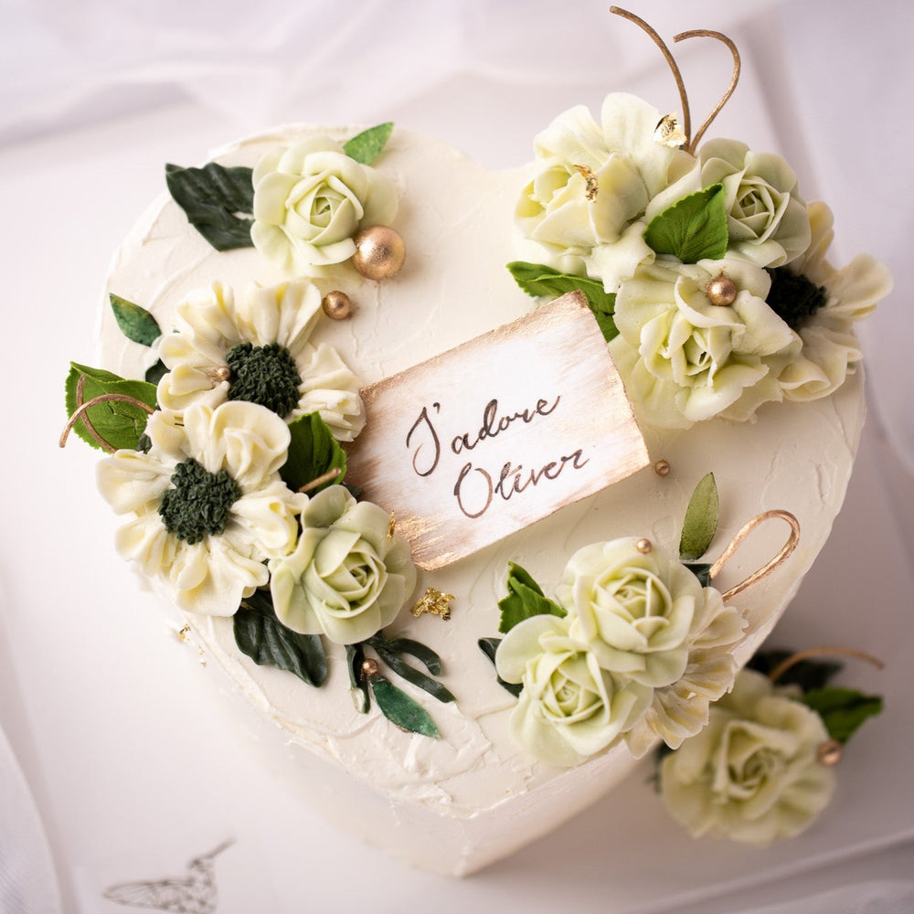 A heart shaped cake with white buttercream base, and light green and ivory buttercream flowers on top. Gold pearls and gold leaves are scattered around the cake delicately. There is a wafer paper note in the middle of the heart that says 