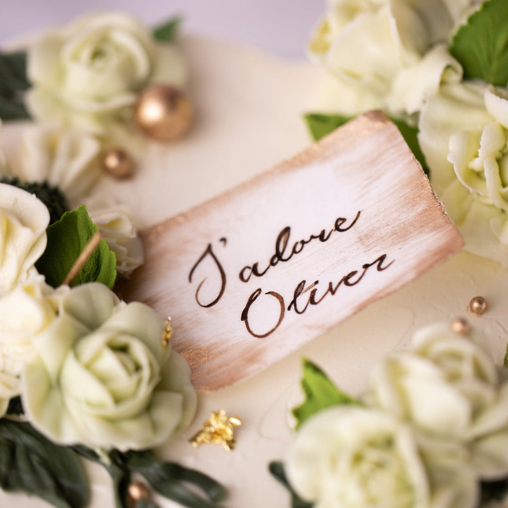 A heart shaped cake with white buttercream base, and light green and ivory buttercream flowers on top. Gold pearls and gold leaves are scattered around the cake delicately. There is a wafer paper note in the middle of the heart that says "J'adore Oliver".