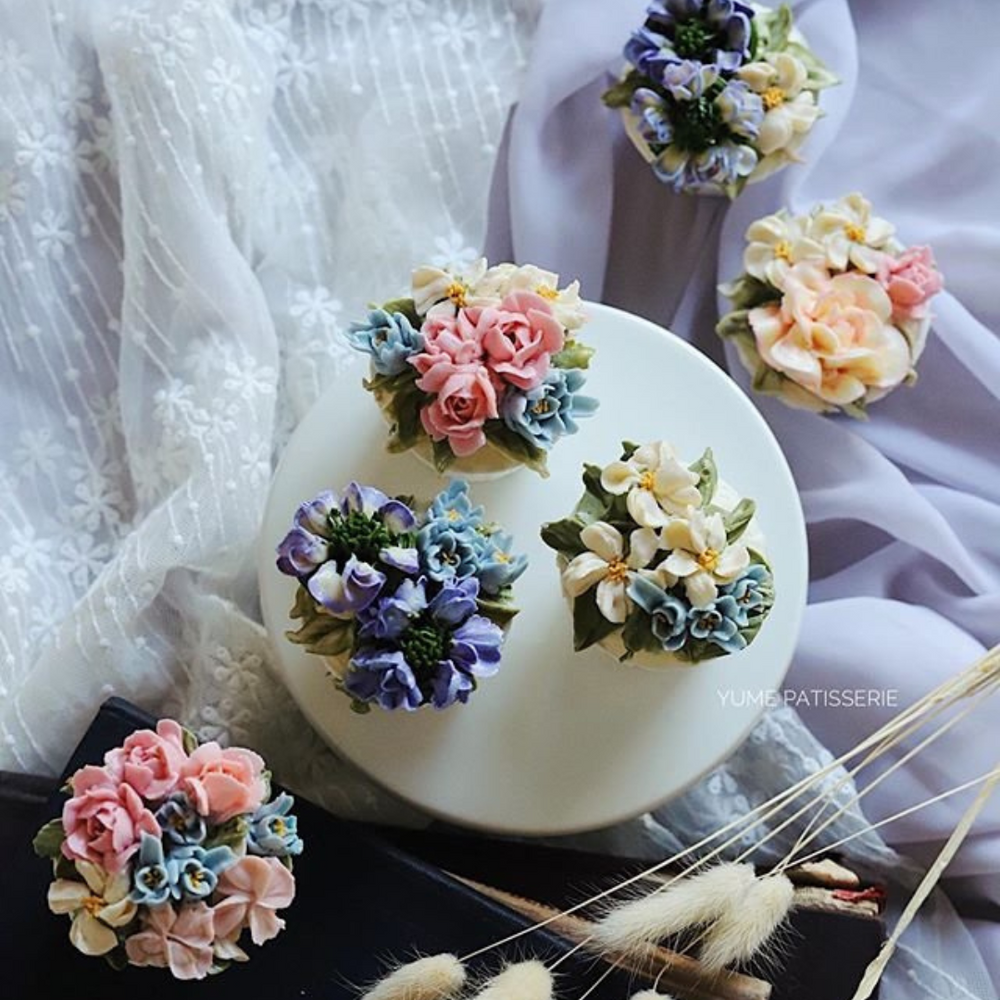 Five cupcakes with assorted hand piped buttercream flowers in various colours. There are three cupcakes in the middle: one with blue flowers, ine with white flowers, and one with pink flowers.