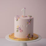 Light pink buttercream cake with little palette knife painted flowers on top and the sides of the cake. The palette knife buttercream flowers are purple, pink and yellow. There is a fondant number One on top of the cake, and the word "flea" in fondant on the front of the cake.