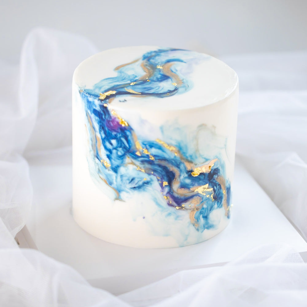 A white fondant cake, with swirls of blue and purple marbled into it, resembling an ocean wave. There are also hand painted swirls of gold on the cake for contrast.