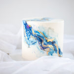 A white fondant cake, with swirls of blue and purple marbled into it, resembling an ocean wave. There are also hand painted swirls of gold on the cake for contrast.