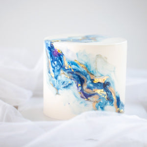 
                  
                    Load image into Gallery viewer, A white fondant cake, with swirls of blue and purple marbled into it, resembling an ocean wave. There are also hand painted swirls of gold on the cake for contrast.
                  
                