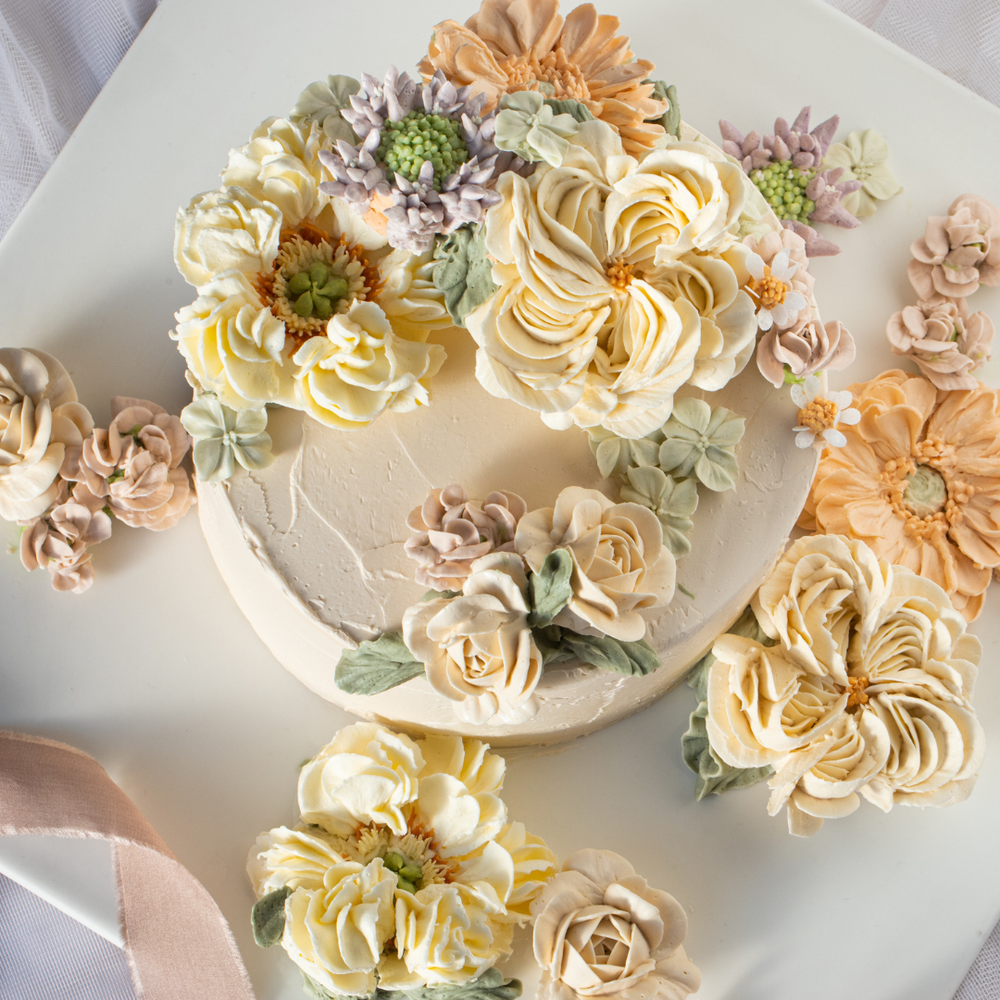 Light ivory buttercream cake with numerous buttercream flowers in white, orange, purple and light green on top. The flowers are 3D and look incredibly realistic.