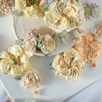 Light ivory buttercream cake with numerous buttercream flowers in white, orange, purple and light green on top. The flowers are 3D and look incredibly realistic.