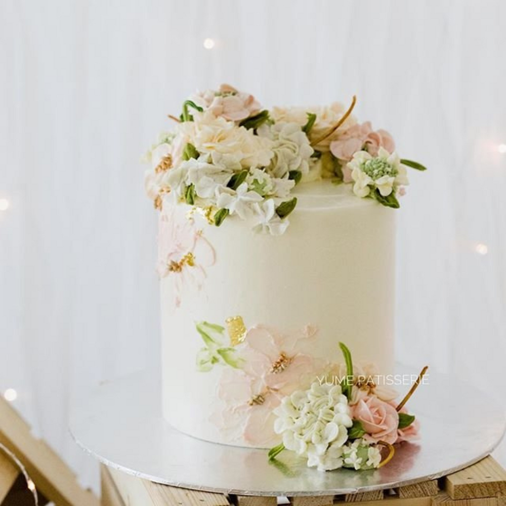 An ivory buttercream cake with white and pink buttercream flowers on top and around it. The cake has hand painted buttercream flowers on the side as well.