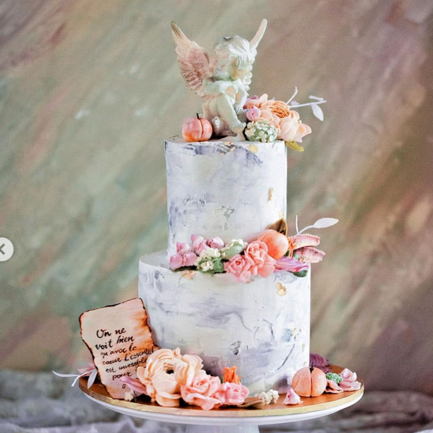 A two tier white and grey marbled buttercream cake. The cake has numerous buttercream flowers in various shades of spring colours. There is a chocolate cherub on top of the cake, and at the base of the cake there is a edible wafer note that resembles a page from a book.