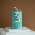 A slim and tall light teal cake that has been sprayed with chocolate spray. There is a white edible wafer sprig with leaves in the center of the cake.