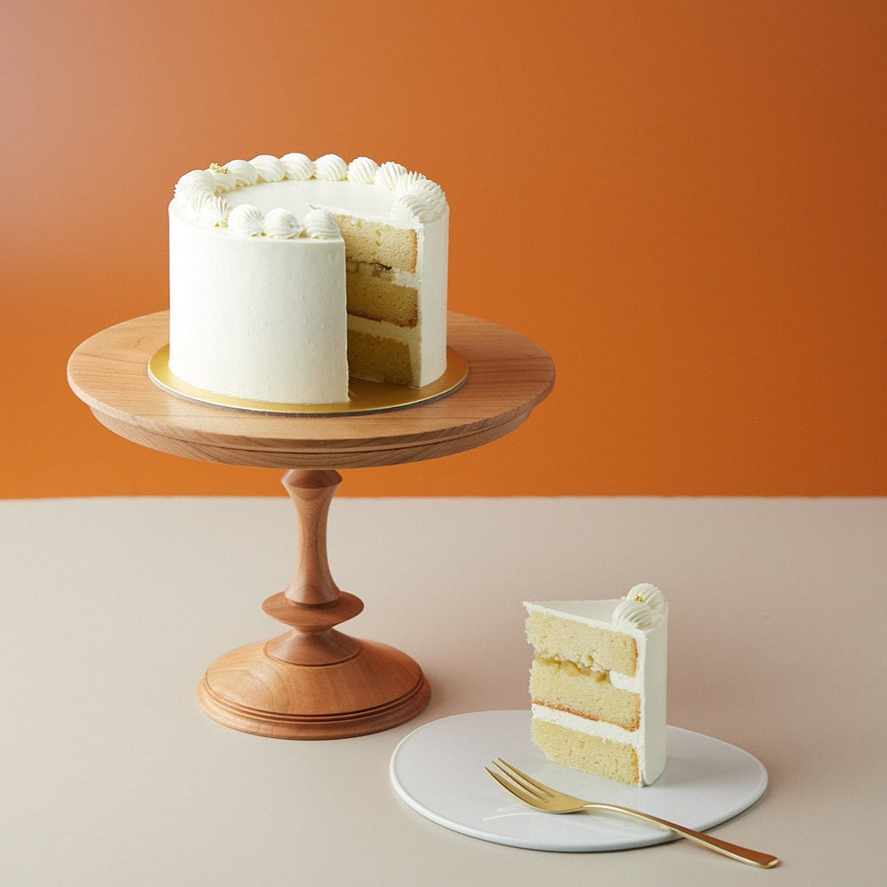 A yuzu apple elderflower buttercream cake, with a slice cut out to show the cross section. The cross section slice is neat and clean, and the sponge is a pale yellow. There is a layer of apple compote between the sponge. The cake is coated in a white elderflower buttercream.
