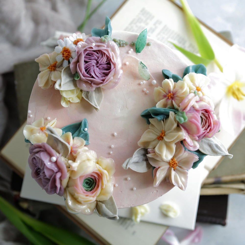 A light pink buttercream cake with white, pink and purple assorted buttercream flowers on top. The cake also has edible sugar pearls delicately scattered on top and around it.