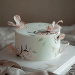 A white fondant cake with hand painted flowers that resemble water colour paintings. The cake has light pink edible wafer flowers on top and beside it, along with specks of gold leaf.