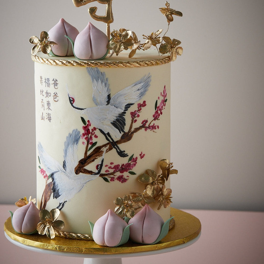 Ivory fondant base with hand painted cranes, and little fondant peaches. Mandarin words are hand painted on the front of the cake. Edible golden flowers adorn the cake.