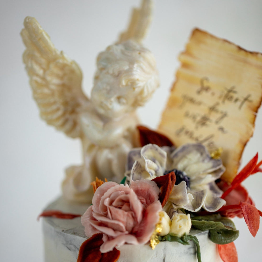 A close up of the white chocolate cherub, and the buttercream flowers. The flowers are pink, purple and white, and there are green and orange edible wafer leaves.