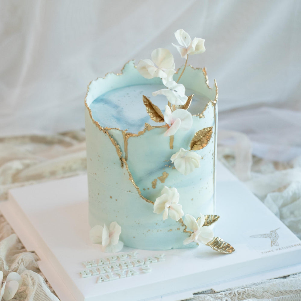 A cake with light blue buttercream carefully wrapped around the side. The buttercream has specks of gold on it, resembling an egg shell. It also has gold luster dust painted on the edges that make the cake pop, along with some edible wafer flowers and gold leaves delicately placed around the cake.