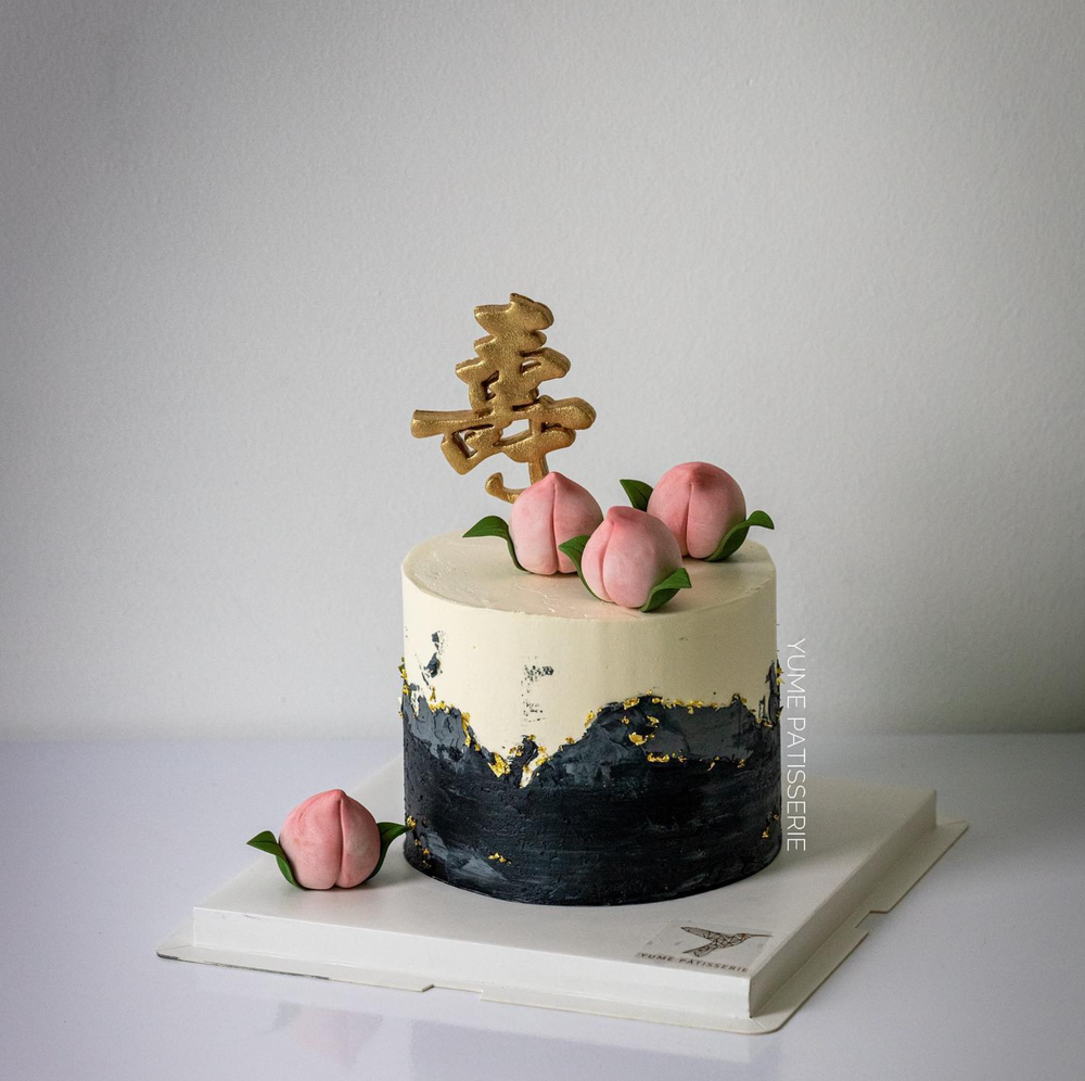 Elegant, clean and modern longevity peach (shou tao) birthday cake Singapore. The cake has a white buttercream base, and the bottom half is dark blue. There are gold accents on the cake, with little peach fondant toppers.