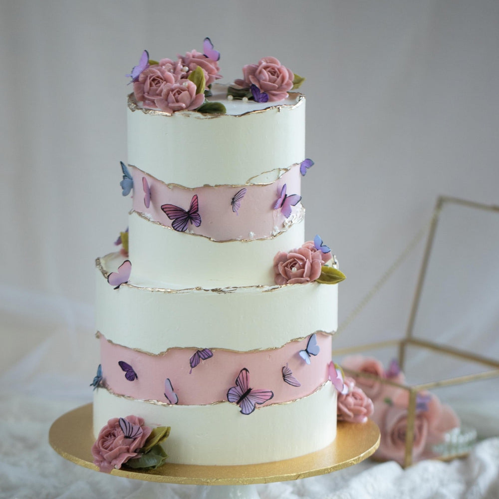 A two tier cake with a white buttercream base adn pink faultline middle. The edges of the faultline are painted with gold luster dust. The middle of the pink faultlines have numerous purple and pink edible wafer butterflies in different sizes. The cake also has multiple pink buttercream roses.