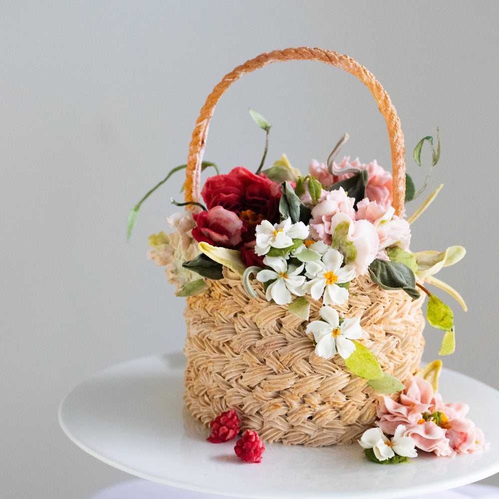 A cake that has been decorated to look like a flower basket. The outside has light brown buttercream that has been piped to resemble a woven basket, and it has assorted buttercream flowers in red, pink and white spilling out of it. It is a realistic flower basket cake.