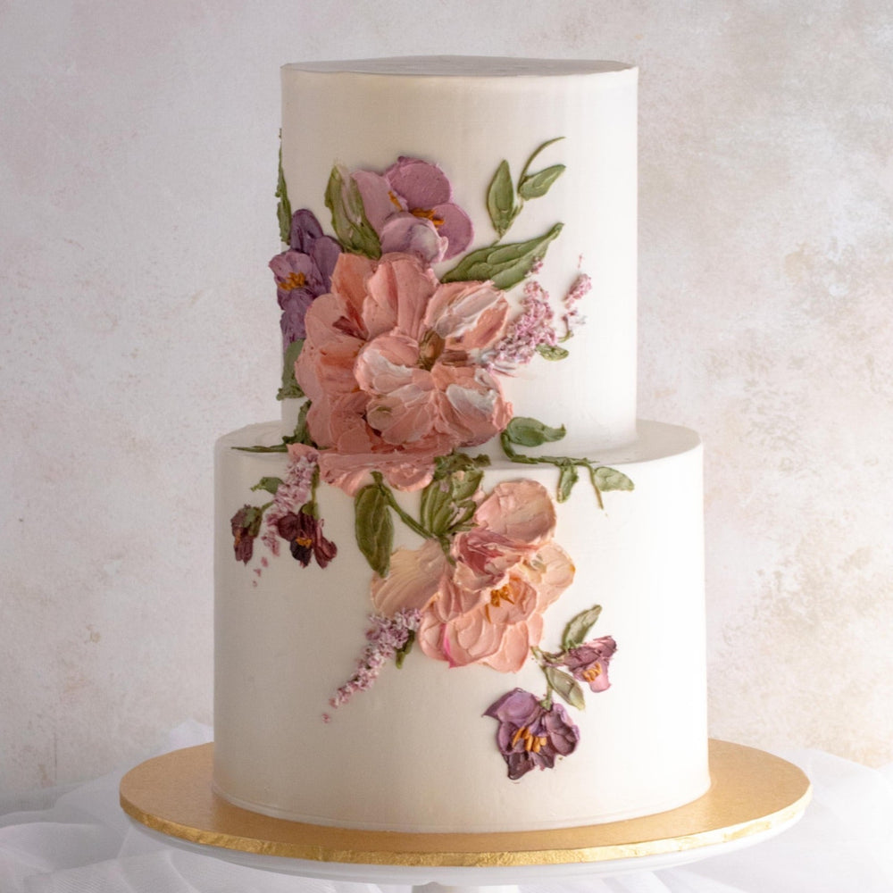 A two tier cake with a white buttercream base, and pink and purple buttercream palette knife flowers on the side of the cake. The flowers are large and detailed, and resemble an oil painting.