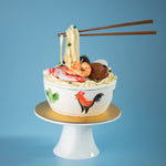 A cake that has been decorated to look like a bowl of noodles. There are fondant shrimp, sliced meat, abalone and spring onions on top of the noodles. Floating above in the :anti gravity" style is a pair of chopsticks also made of fondant, holding into the fondant noodles.