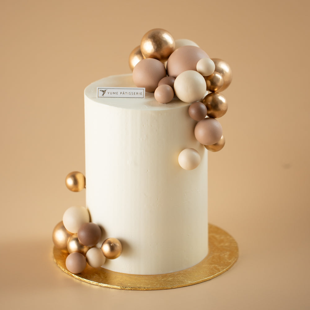 A tall and slim cake with a white buttercream base. The cake has numerous shiny and matte round ornaments in gold and nude colours on the top and bottom of the cake.