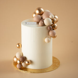 
                  
                    Load image into Gallery viewer, A tall and slim cake with a white buttercream base. The cake has numerous shiny and matte round ornaments in gold and nude colours on the top and bottom of the cake.
                  
                