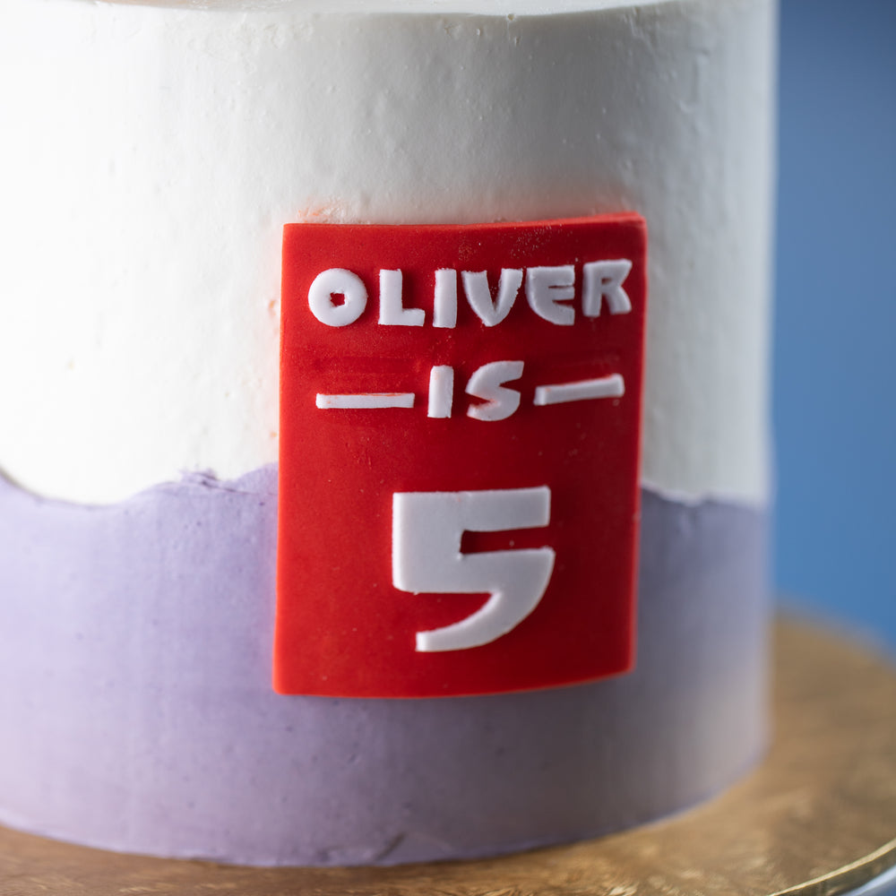 
                  
                    Load image into Gallery viewer, A close up on the red fondant rectangle in the middle of the cake. It says &amp;quot;Oliver is 5&amp;quot; in the Big Hero 6 movie font.
                  
                