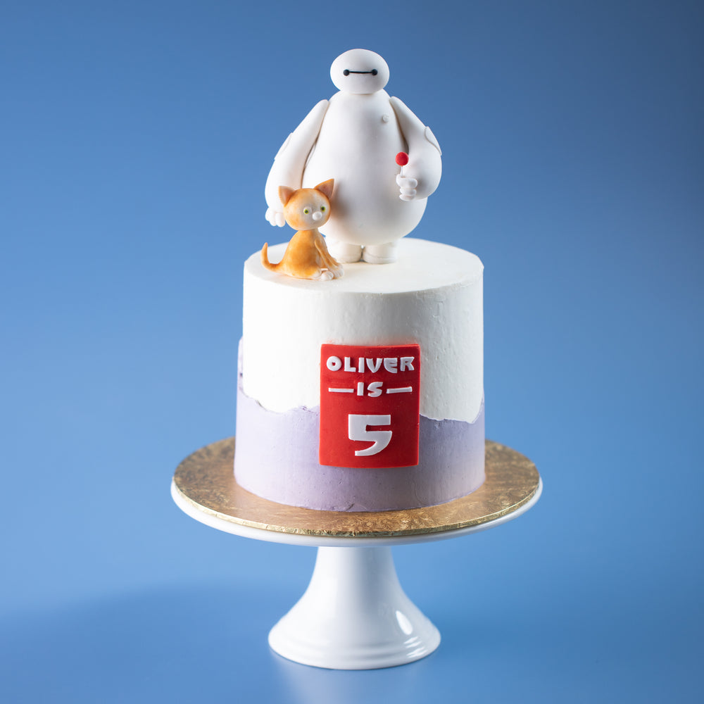 A cake with a white buttercream base, with a jagged light purple buttercream at the bottom half of the cake. The cake has a red fondant rectangle in the middle with the Big Hero 6 font. The cake is in Baymax's armour colours. At the top of the cake there is a 3D fondant Baymax holding his signature red lolipop. To his right, there is an orange cat sitting by his feet.